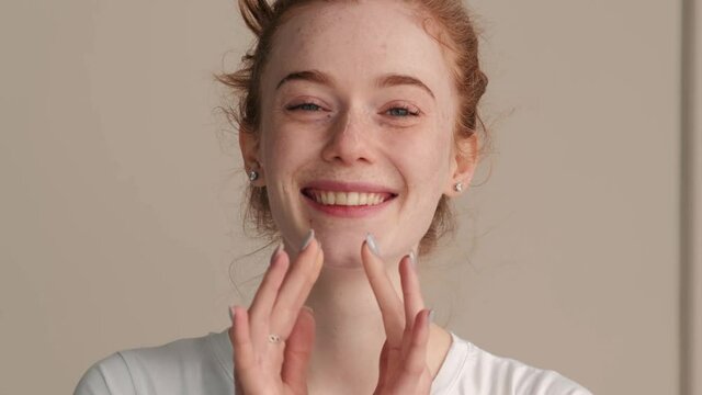 Adorable woman with freckles and red hair is smiling at camera while applying cream on her face