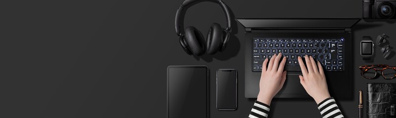 黒いテーブルの上でノートパソコンにタイピングしている人物のイメージ。卓上にあるスマートフォン、タブレット、スマートウォッチなどダークトーンの物によるイメージ。真上からのアングル