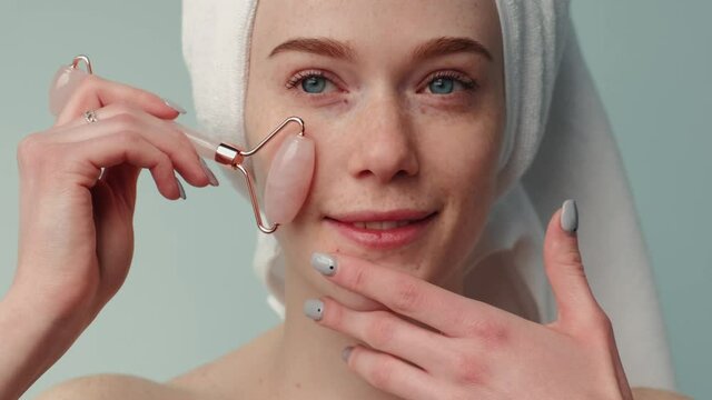 Freckled woman with ginger hair is massaging her face with a derma roller smiling at camera