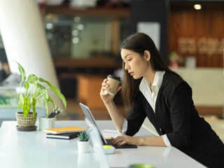 Female office worker drinking coffee while working with digital tablet in office room