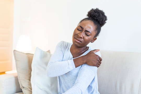 African woman sitting on couch take break touches her shoulder suffers from painful feelings ache caused by poor wrong posture, sedentary work, sitting at laptop for long period concept image