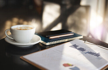 Workplace on desk, close-up white coffee cup with paperwork of investment and mobile phone, pen on book
