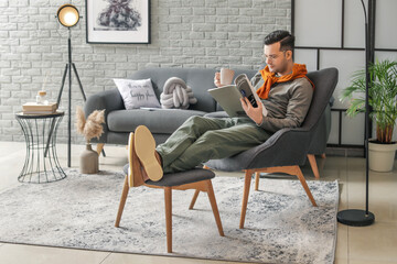 Young man reading magazine and drinking tea while sitting in armchair at home