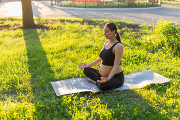 Peaceful young positive pregnant woman in gymnastic suit does yoga and meditate sitting on mat on green grass on sunny warm summer day. Concept of preparation for childbirth and positive attitude