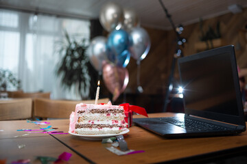 A large piece of cake and a candle stands on the table near the laptop. Birthday celebration.
