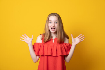 Cute girl raises palms from joy, happy to receive awesome present from someone, shouts loudly, dressed in summer dress, isolated on yellow background. Excited teen girl yells.