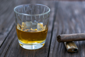 A glass of whiskey stands on a vintage wooden table. A cigar and a lighter lie nearby 