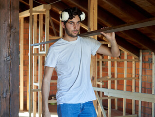 Young carpenter with beard and white t-shirt carrying a wooden slat in a house under construction man with hard hats on his head working on a wood work.