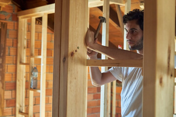 Young carpenter with a beard and a white t-shirt driving nails with a hammer in a house under construction, with a lot of wood in the background. Man with hard hats on his head working