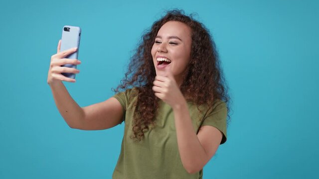Funny young african american woman in green t-shirt isolated on blue background studio. People lifestyle concept. Doing selfie shot on mobile phone victory sign thumb up blinking blowing send air kiss