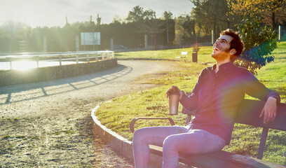 young man in blue shirt having coffee on a bench laughing while having coffee, with the sunset in the background. teenager having coffee in the park. taking a break in the park