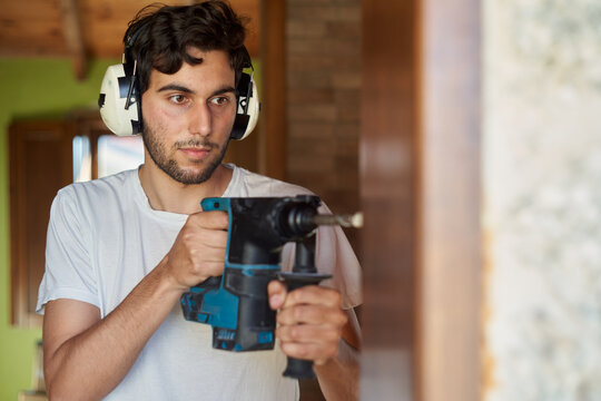 Young Carpenter, With Hard Hat, Beard And White T-shirt With Blue Drill Making A Hole In The Wall. Man Drilling The Frame. Teenager On His First Job