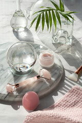 Gua Sha massager and pink quarz face roller on marble stone background. Face care products, glass crystal ball and exotic palm leaves. Towel,moisturizing cream, skincare tonic, essential oil, serum.