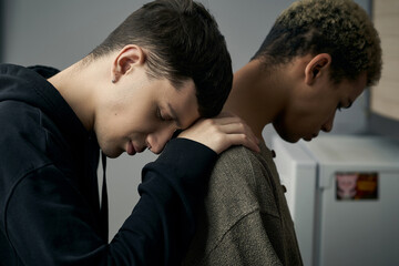 Staged photo illustrates problems and conflicts in gay couple relationships. Young black haired man is hugging swarthy man from behind in the kitchen.