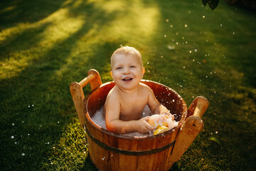 baby splashes in a basin in nature on a green tvava, at sunset. Good mood, child's smile, happiness from swimming in water, water splashes fly, good summer day