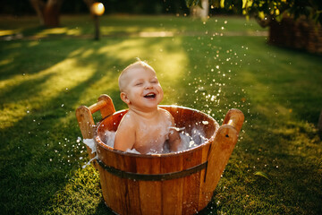 baby splashes in a basin in nature on a green tvava, at sunset. Good mood, child's smile, happiness from swimming in water, water splashes fly, good summer day