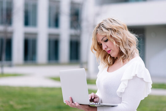 horizontal image of a 30 year old woman using a laptop computer against a blurred background of modern buildings. The blonde-haired executive communicates in an online business meeting.