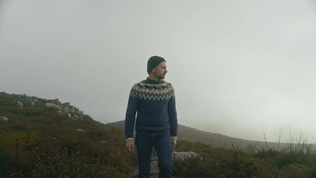Cinematic moody man in traditional wool sweater walk in empty field, high in mountains in fog. Lonely solo adventure trip, slow motion of guy in warm outfit hike in epic scenery on cloudy day