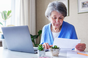 Elderly woman reads a bank loan notice or letter - Grandmother works from home with laptop and paperwork - Planning budget and retirement benefits and managing finances - obrazy, fototapety, plakaty