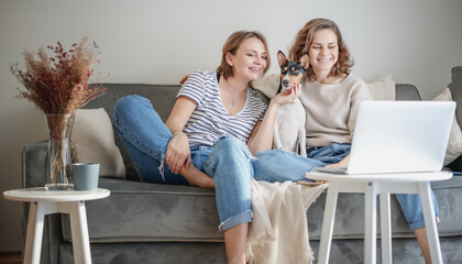 Beautiful happy lesbian couple sitting on sofa at home with pet dog bassengi online chatting using laptop