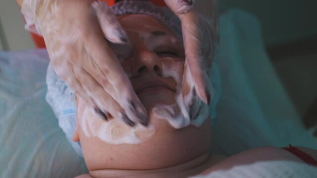 CU: relaxed young lady with overweight undergoes face cleaning procedure with foamy mask by skilled beautician in cosmetology clinic closeup