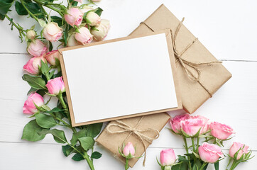 Greeting card mockup with fresh roses and gift boxes on white wooden background
