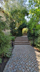 Park landscape. Path through arched gateway to English garden with retro streetlights by sides. Stone paved stairs in park with hedge arch