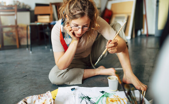 Creative female artist sitting on the floor in the art studio and painting on paper with a paintbrush. A woman student painter in eyeglasses painting with watercolors in her workshop.