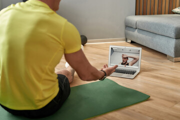 Virtual fitness class. Rear view of a mature fitness coach in sportswear sitting on yoga mat at home and conducting online training