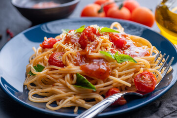 Tasty appetizing classic italian spaghetti pasta with tomato sauce, cheese parmesan and basil on plate and ingredients for cooking pasta on dark table.