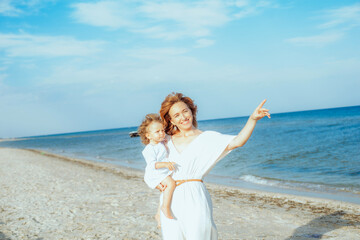 Fototapeta na wymiar Mom is holding and smiling a little daughter 1 year old with red curly hair in light clothes in the summer on the beach. Happy childhood. Travel and family vacations 