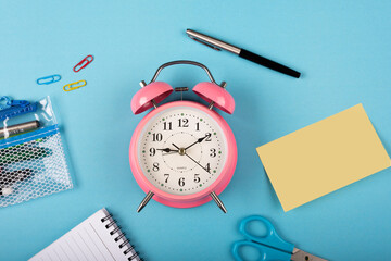 Table clock with pen and Scissors and sticky notes on a blue background flat lay shot