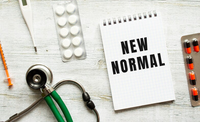 NEW NORMAL is written in a notebook on a white table next to pills and a stethoscope.