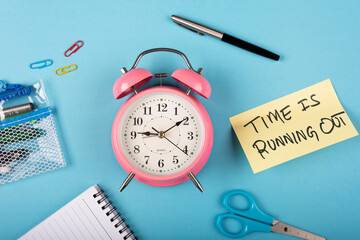 A pink color table clock with Time is running out words Written on a yellow sticky note and Pen with other elements on a blue background.