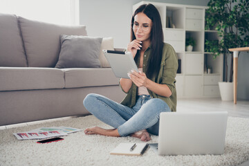 Full size photo of attractive positive lady sit on floor barefoot hand on chin look minded tablet working home indoors