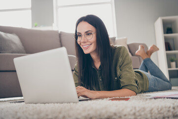 Full size photo of pretty positive lady lying on floor beaming smile look laptop have good mood working home indoors