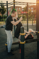 The man the trainer observes the girl performing the exercise on the press of the urban sports ground on an inclined bench
