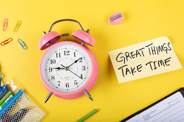 A table clock with Great things take time words Written on a sticky note and Sharpener with other elements on a Yellow background flat lay shot.