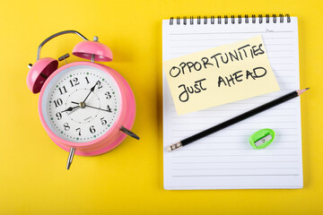 A table clock with Opportunities just ahead words Written on a sticky note with notepad pencil and other elements on a Yellow background.