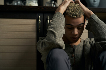 Obraz premium Staged photo illustrates problems and conflicts in gay couple relationships. Swarthy man in gray cardigan is sitting on the floor near the kitchen base cabinet. Wistful man grabbed his head.