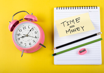 A table clock with Time equal to money words Written on a sticky note with notepad pencil and other elements on a Yellow background.