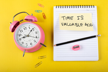  A table clock with Time is valuable acid words Written on a sticky note with notepad pencil and other elements on a Yellow background.