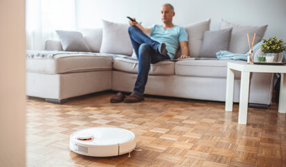 Man Relaxing On Sofa With Robotic Vacuum Cleaner On Hardwood Floor. Cleaning concept - automatic robotic hoover clean the room while man relaxing, close up