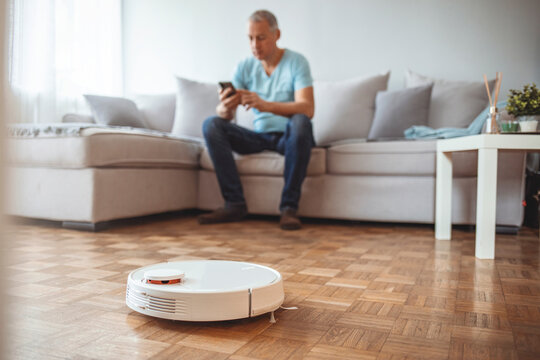Young man using automatic vacuum cleaner to clean the floor, controling machine housework robot. Happy Man Lying On Sofa Operating Robotic Vacuum Cleaner With Remote Control