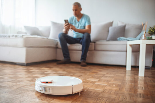 Young man using automatic vacuum cleaner to clean the floor, controling machine housework robot. Happy Man Lying On Sofa Operating Robotic Vacuum Cleaner With Remote Control