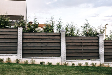 Horizontal tiered sections of brown wooden boards fence and white brick pillars. Live plantings....