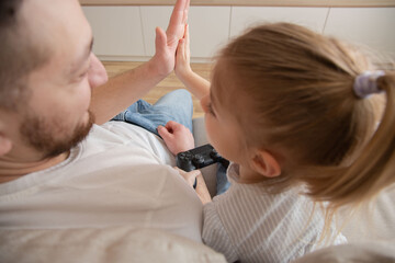 Fototapeta premium young man father giving high five to happy little child daughter after finishing video game. Joyful two generations family celebrating successful win, playing together in living room. Back view.
