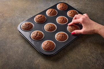 Woman's hand with a manicure pulls out a chocolate cupcake from baking dish on wooden background, close up