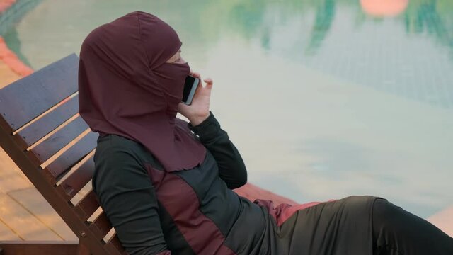 Muslim woman talking on phone near pool sitting on deckchair.