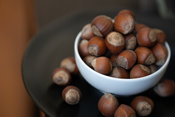 Heap, pile of brown nuts, hazelnuts lies in a small white plate and a black plate on table on blurry warm background. Close-up.